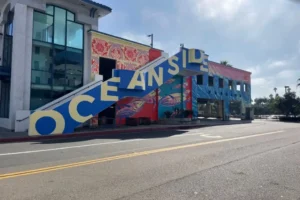 A colorful building features a large staircase with the word OCEANSIDE painted in blue and yellow letters. Murals of fish and waves decorate the walls, and the street in front is empty under a partly cloudy sky.
